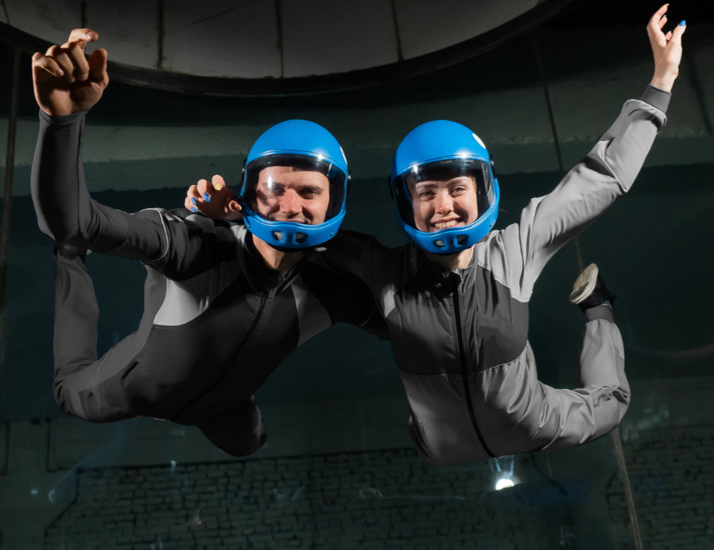 Two smiling indoor skydivers in blue helmets float in a wind tunnel, arms raised against a dark background.